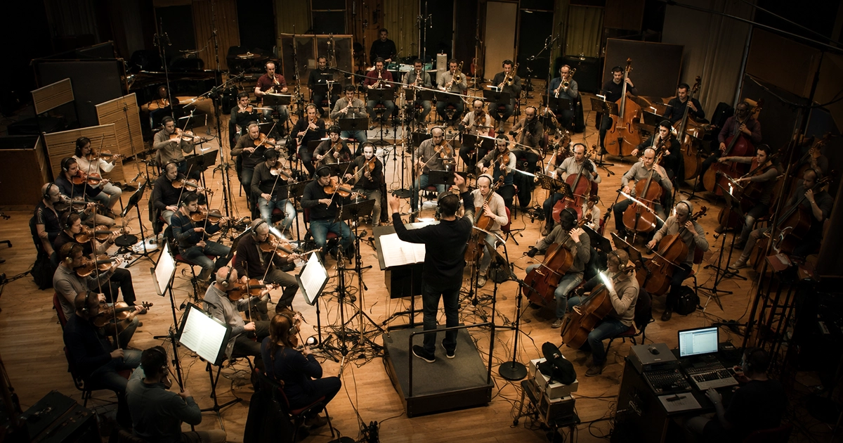 A full orchestra recording in a studio, viewed from a high overhead angle, with the conductor mid-gesture surrounded by strings, cellos and double basses