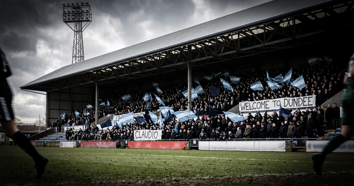 A packed stand at a Scottish football ground under a stormy sky, fans waving Argentine and navy flags and holding banners reading 'Claudio' and 'Welcome to Dundee'